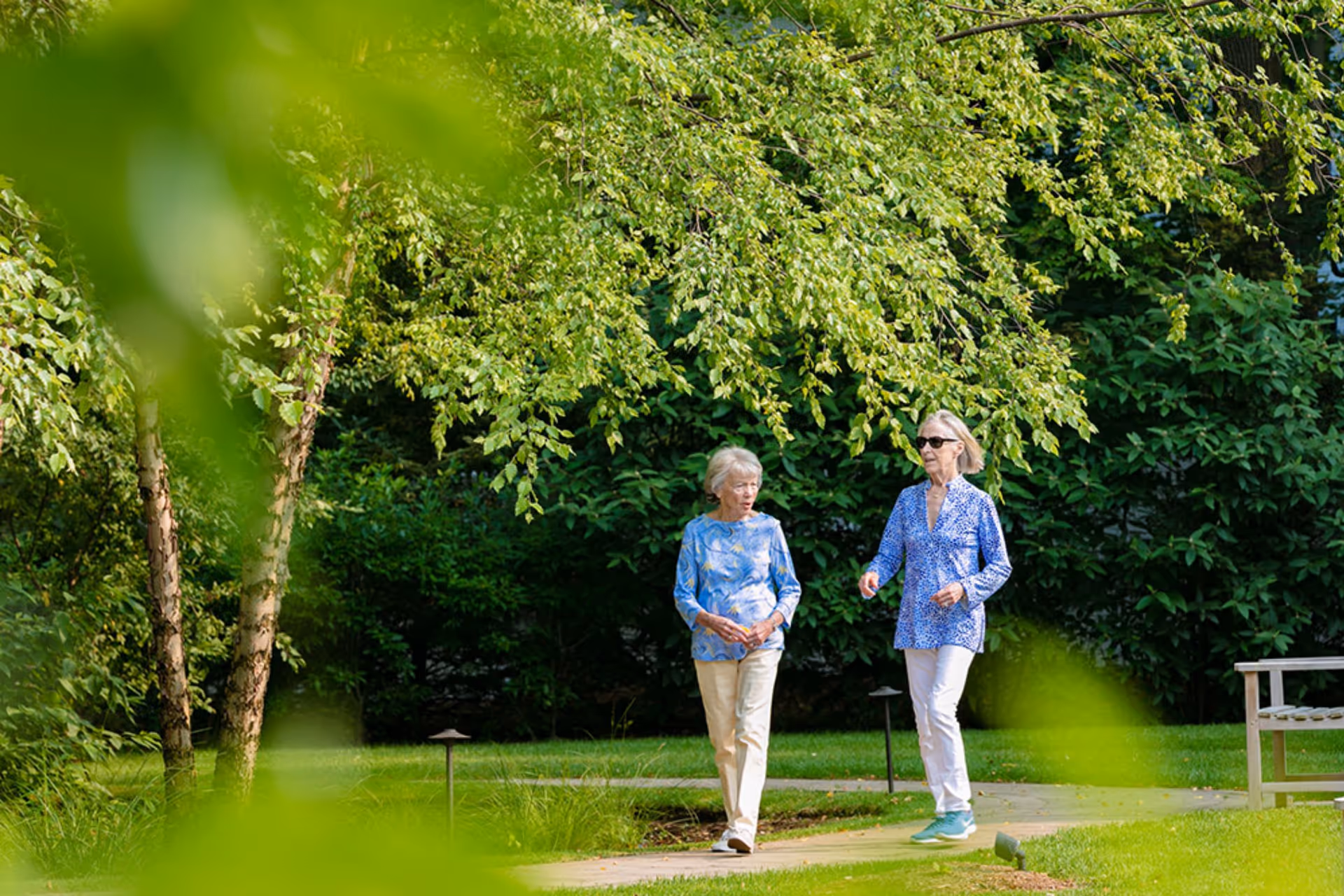 Two elderly women walking and talking on a paved path in a lush green garden with trees and bushes surrounding them. One woman is wearing a blue floral top and beige pants, and the other is wearing a blue patterned shirt with white pants and sunglasses. A wooden bench is visible on the right side of the path.