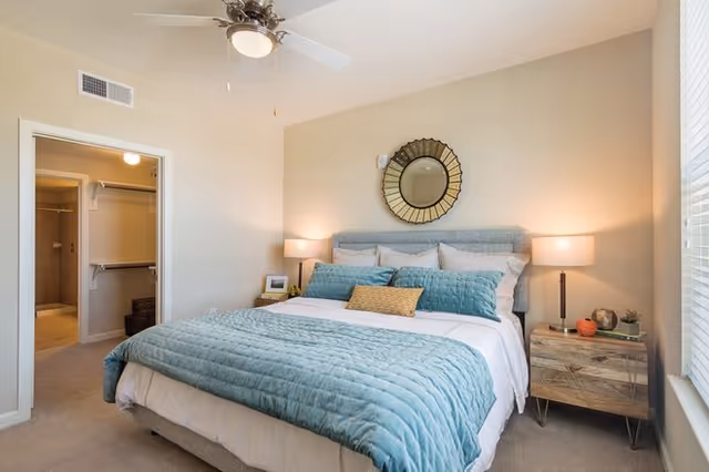 Sunlit bedroom with a teal-covered bed, two nightstands, a decorative round mirror above the headboard, and a ceiling fan.