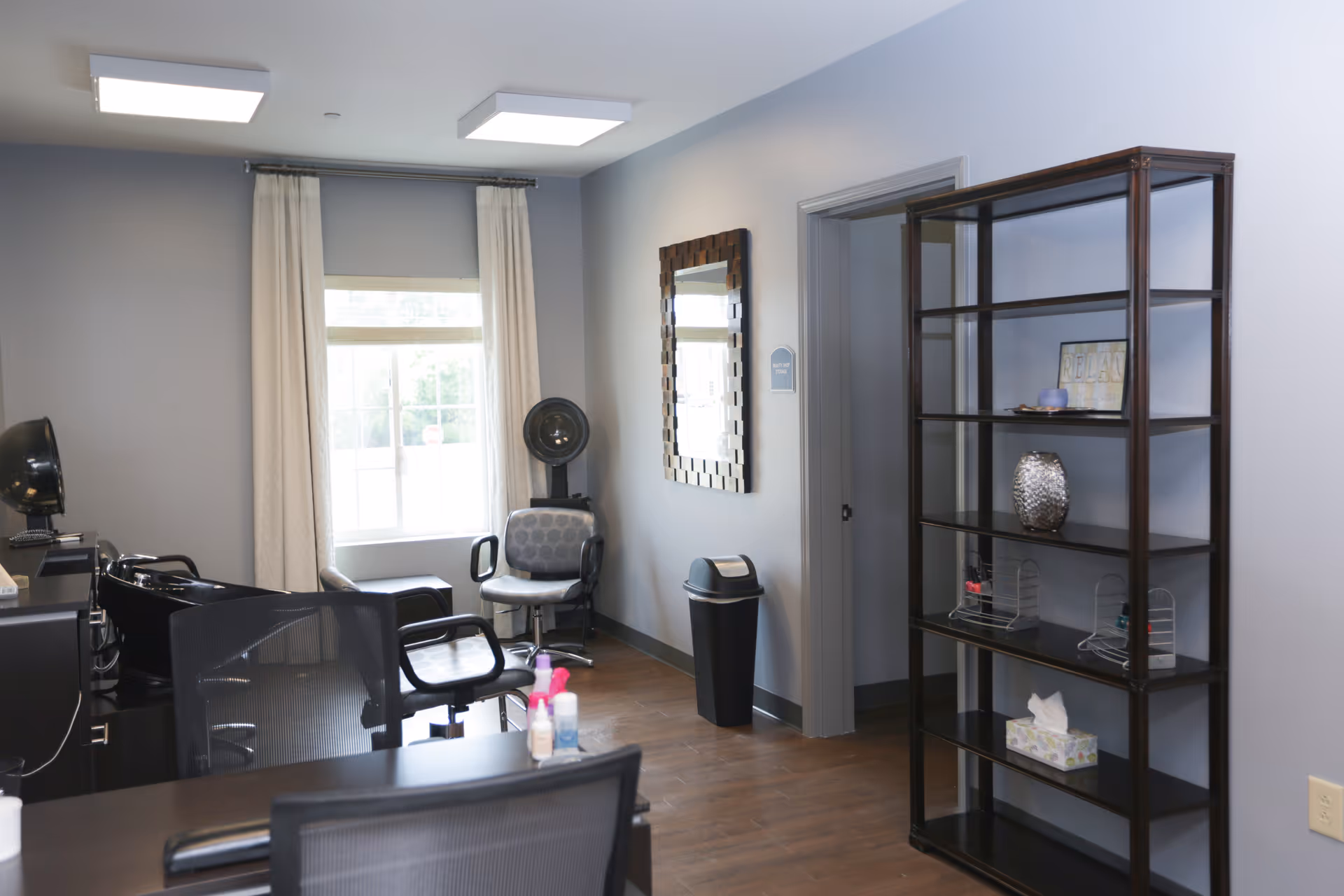 Interior view of a salon area in an assisted living facility with salon chairs, a hair washing station, a window with curtains, a decorative mirror on the wall, a trash bin, and a tall shelving unit with various items including a tissue box and decorative objects.