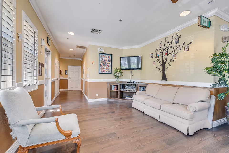 A bright senior living lounge with a beige sofa, armchair, wall-mounted TV, and a decorative photo tree on the wall.
