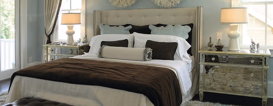 A neatly made bed with a tufted beige headboard, white sheets, multiple pillows in white, black, and light blue, and a brown blanket. On each side of the bed are mirrored nightstands with table lamps and decorative items. Light enters the room through a window and a glass door with curtains.