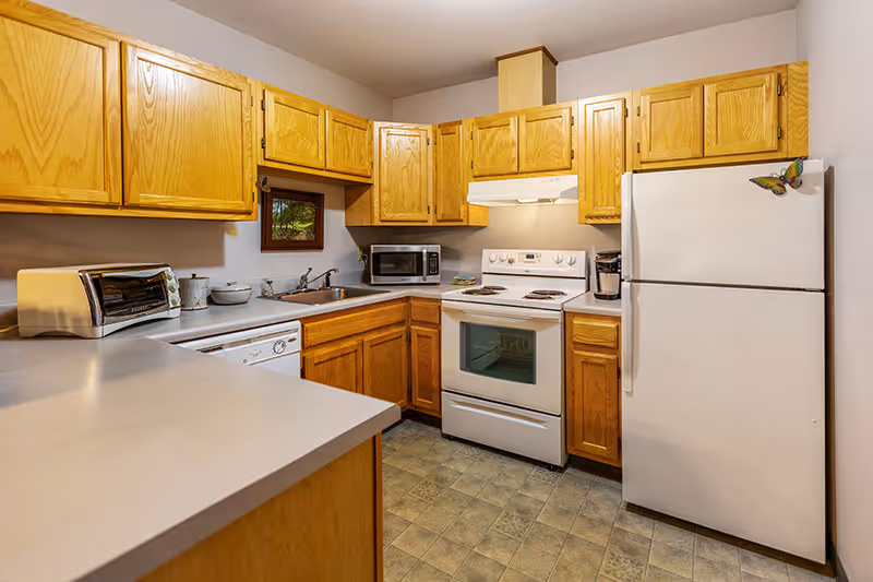 A compact kitchen with wooden cabinets, a white refrigerator with a butterfly magnet, a white electric stove with oven, a microwave, a toaster, a coffee maker, and a dishwasher. The countertops are light-colored, and there is a small window above the sink.