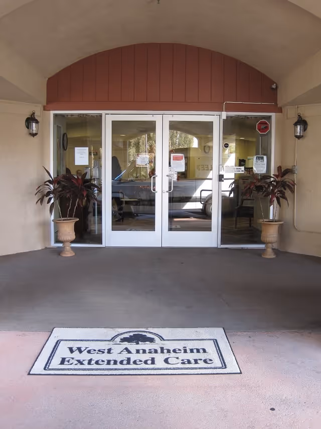 Entrance to a building with double glass doors under an arched roof. Two potted plants flank the doors, and a doormat on the ground reads 'West Anaheim Extended Care'.