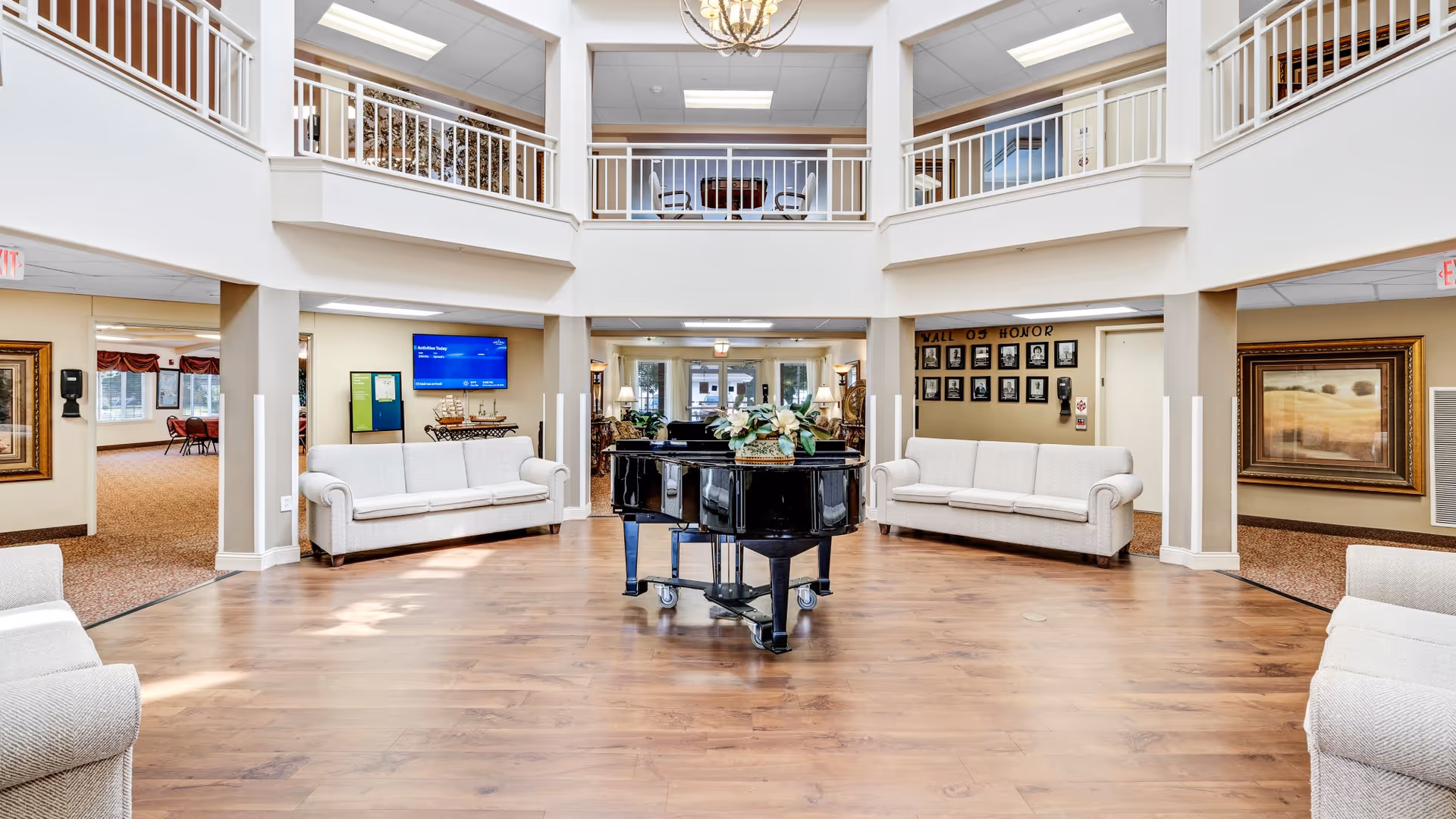 Spacious senior living facility common area with a black grand piano in the center, surrounded by four light-colored sofas. The room has a high ceiling with a chandelier, a second-floor balcony with white railings, and wood flooring. There are framed pictures on the walls, a TV screen displaying activities, and a 'Wall of Honor' with photos on the right side.