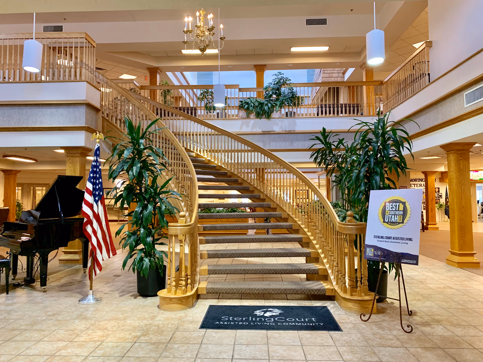 Interior view of Sterling Court assisted living community featuring a grand wooden staircase with carpeted steps, two large potted plants on either side, an American flag, a black grand piano, and a sign that reads 'Best of Southern Utah - Sterling Court Assisted Living'. The area has tiled flooring and a chandelier hanging from the ceiling.