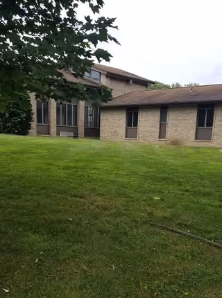 Exterior view of a single-story and two-story building with beige brick walls and several windows, surrounded by a well-maintained green lawn and partially shaded by tree branches.