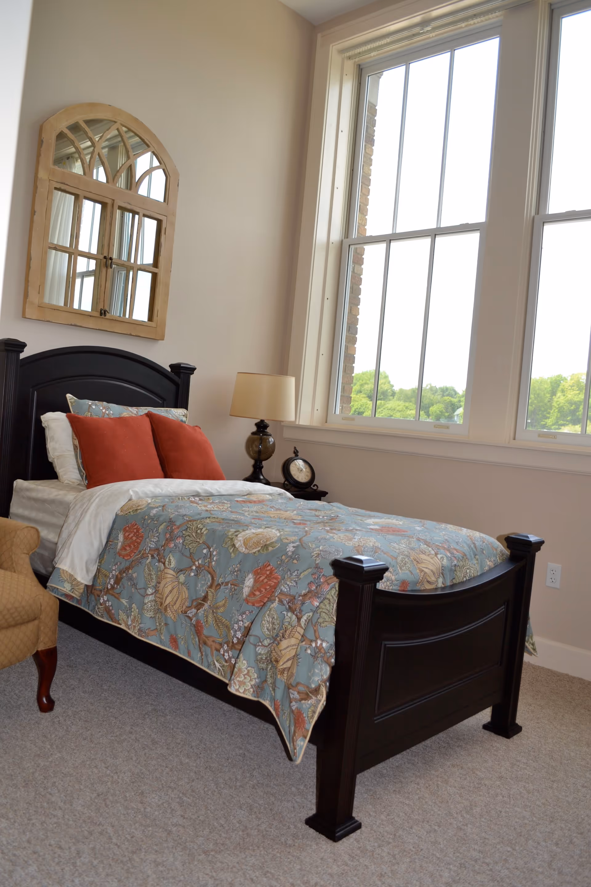 A bedroom with a dark wooden single bed featuring a floral patterned bedspread and two red pillows. Next to the bed is a small table with a lamp and a clock. A large window with white frames lets in natural light, and a decorative wooden-framed mirror hangs on the beige wall above the bed.