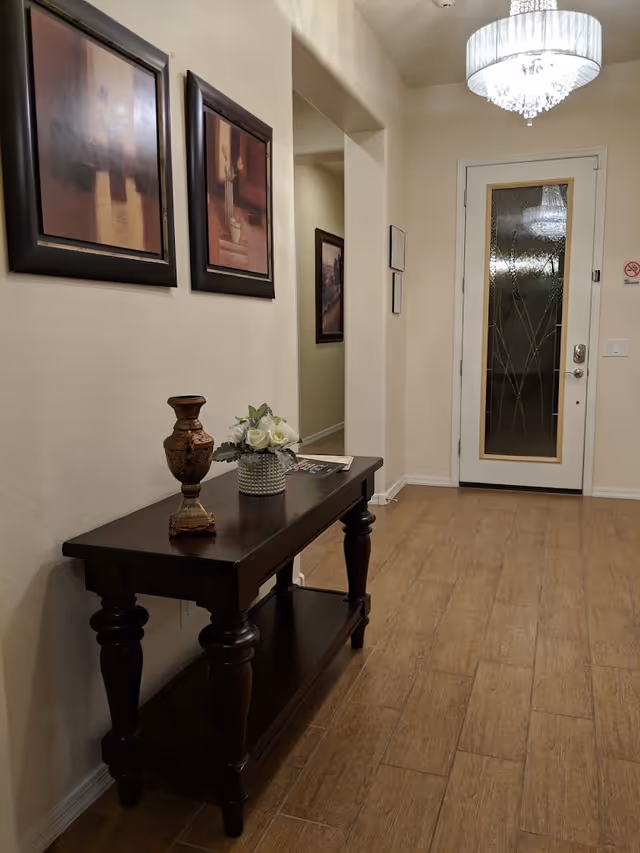 A hallway with wooden flooring and cream-colored walls. On the left side, there is a dark wooden console table with a decorative vase and a flower arrangement. Above the table, two framed abstract paintings hang on the wall. At the end of the hallway, there is a door with a decorative glass panel and a chandelier reflected in the glass.