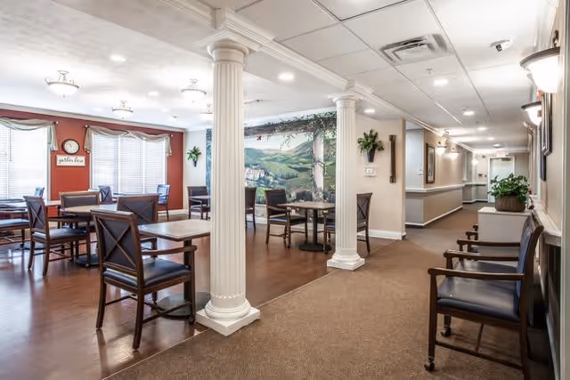 Interior view of a senior living facility dining area with several tables and chairs arranged on a wooden floor. The space features white columns, a mural of a countryside landscape on one wall, and large windows with blinds. Adjacent to the dining area is a carpeted hallway with additional seating and wall-mounted lights.