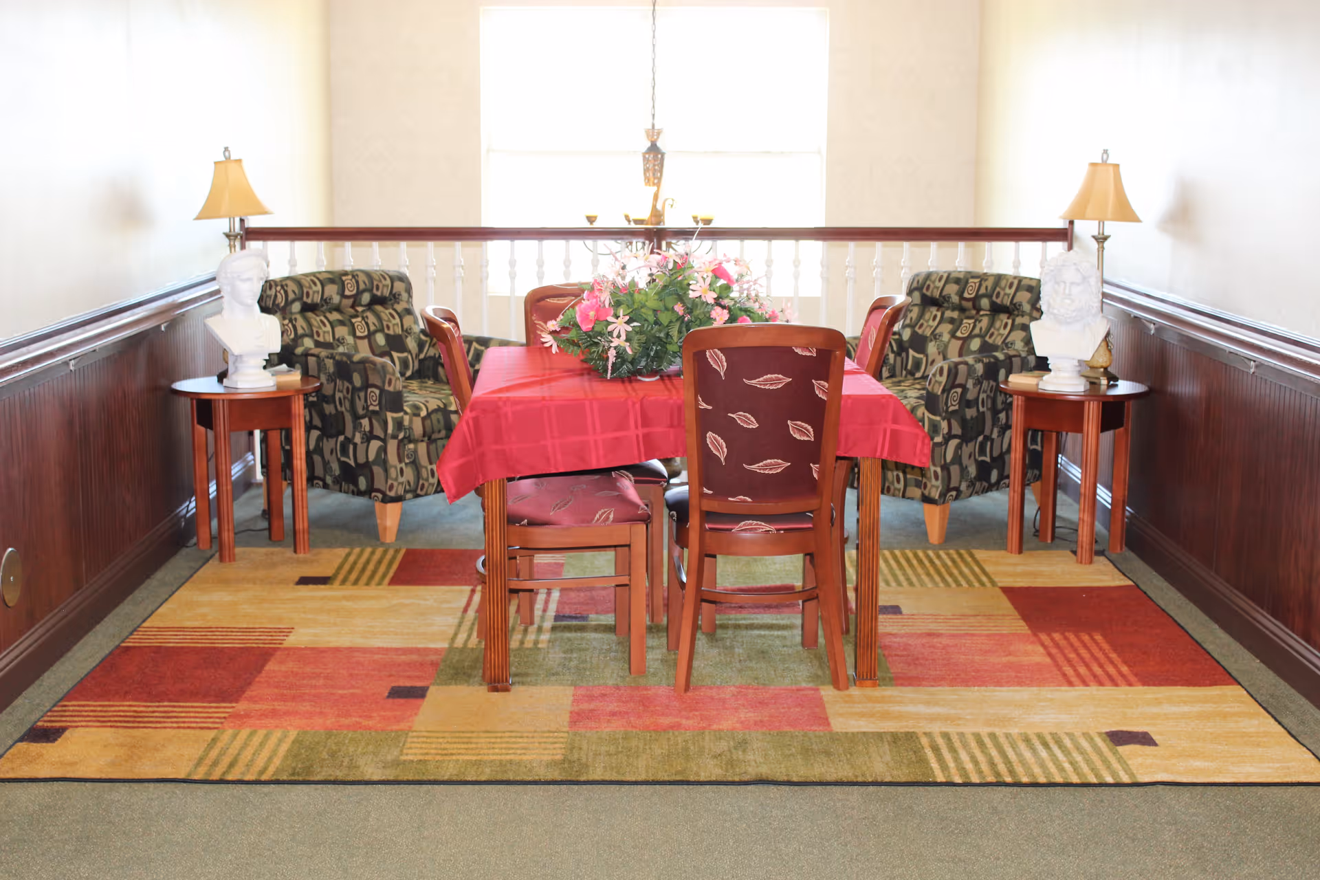A cozy sitting area with a rectangular wooden table covered with a red tablecloth and a floral centerpiece. Four wooden chairs with patterned upholstery surround the table. Two patterned armchairs are positioned against the wall on either side, each next to a small round wooden side table holding a white bust sculpture and a lamp. The floor features a colorful geometric patterned rug, and a large window in the background lets in natural light.