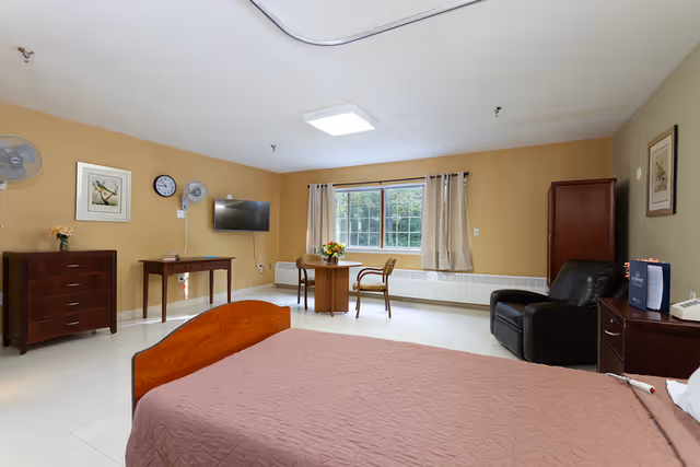 A spacious senior care bedroom with a bed in the foreground, a wall-mounted TV, dresser, armchair, and a small table with chairs by a window.