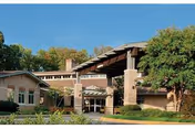 Exterior view of a senior living community building with a covered entrance, surrounded by trees and greenery under a clear sky.