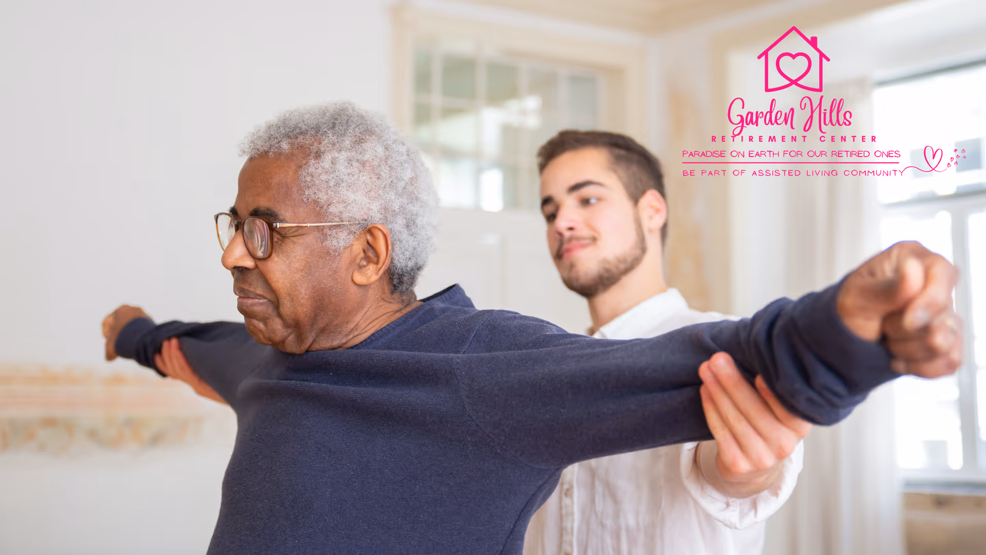 An elderly man with gray hair and glasses is stretching his arms out wide while a younger man assists him with the exercise in a bright room with large windows.