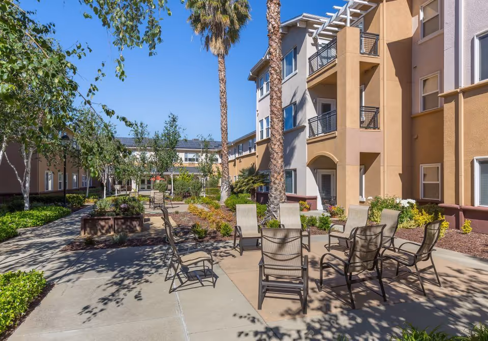 Outdoor courtyard area at Heritage Estate Senior Apartments featuring a paved seating area with multiple chairs arranged in a circle, surrounded by landscaped plants, trees including palm trees, and a multi-story residential building with balconies in the background under a clear blue sky.