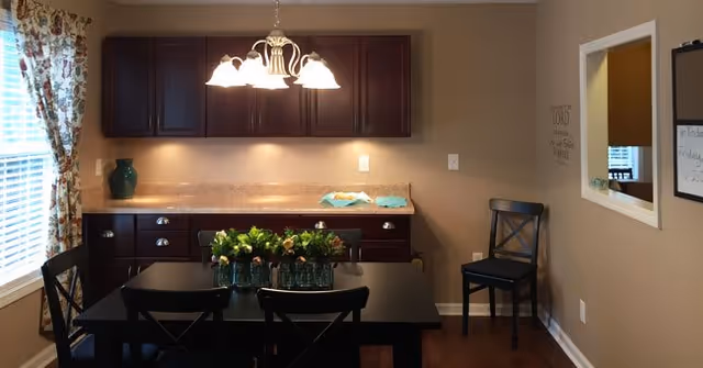 A dining area with a black dining table and four black chairs. On the table are several small vases with flowers. Behind the table is a countertop with dark wooden cabinets above and below. A chandelier with five lights hangs above the table. To the left, there is a window with floral curtains, and on the right wall, there is a mirror and a small whiteboard calendar.