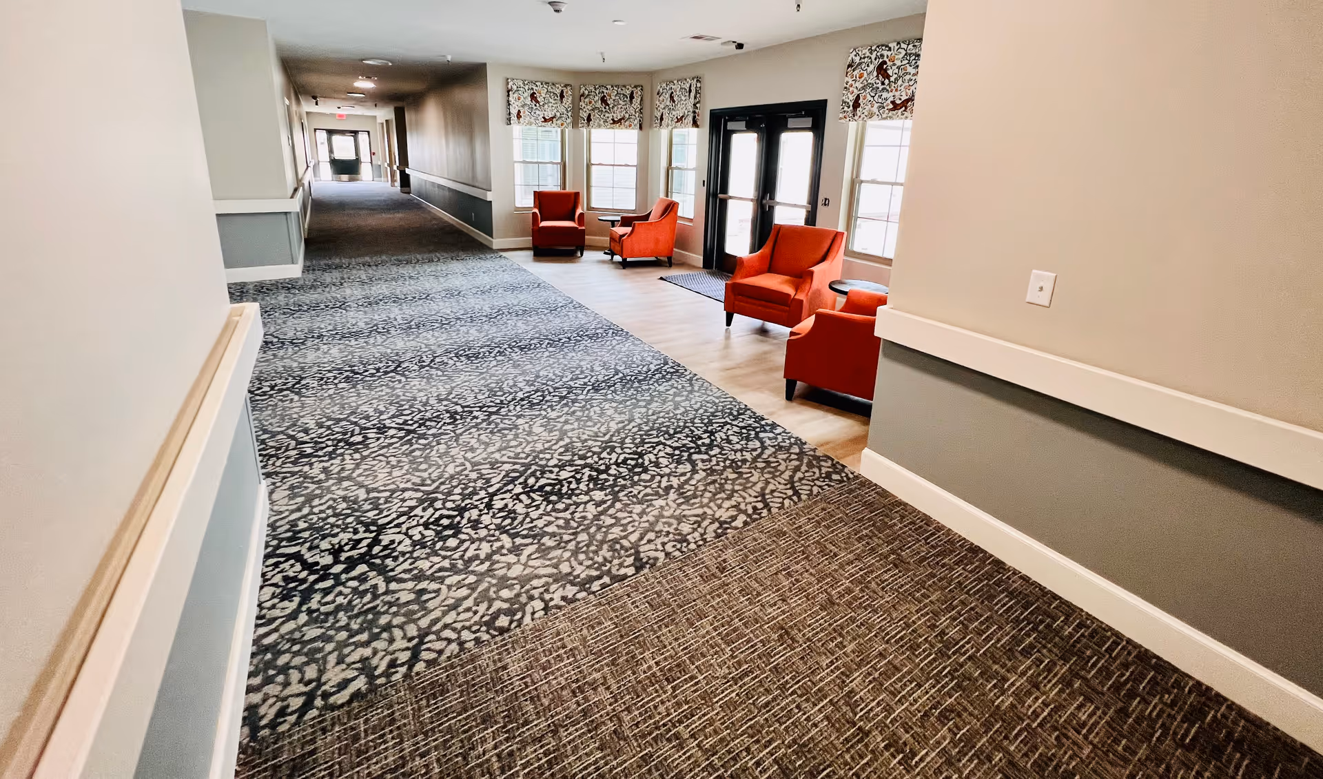 A long hallway in a senior living facility with patterned carpet and beige walls. At the far end near windows and glass doors, there are four red armchairs arranged in pairs with small round tables between them. The hallway is well-lit with natural light coming through the windows.