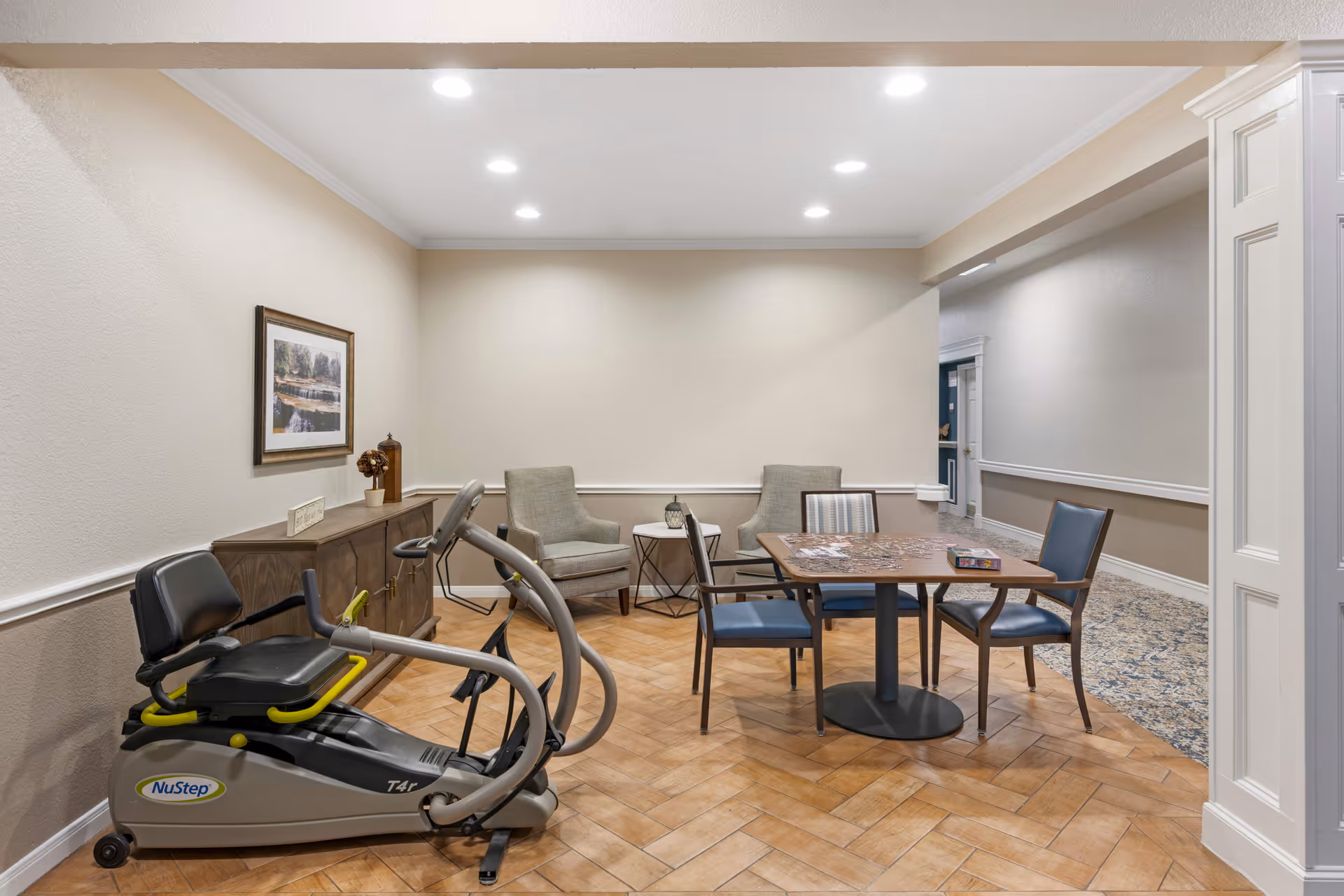 A cozy senior living common area with a NuStep exercise machine on the left, a wooden cabinet with decorative items and a framed picture above it, two upholstered armchairs with a small table between them, and a square table with four chairs. The table has a partially completed puzzle and a puzzle box on it. The room has recessed ceiling lights and a mix of wood and carpet flooring.