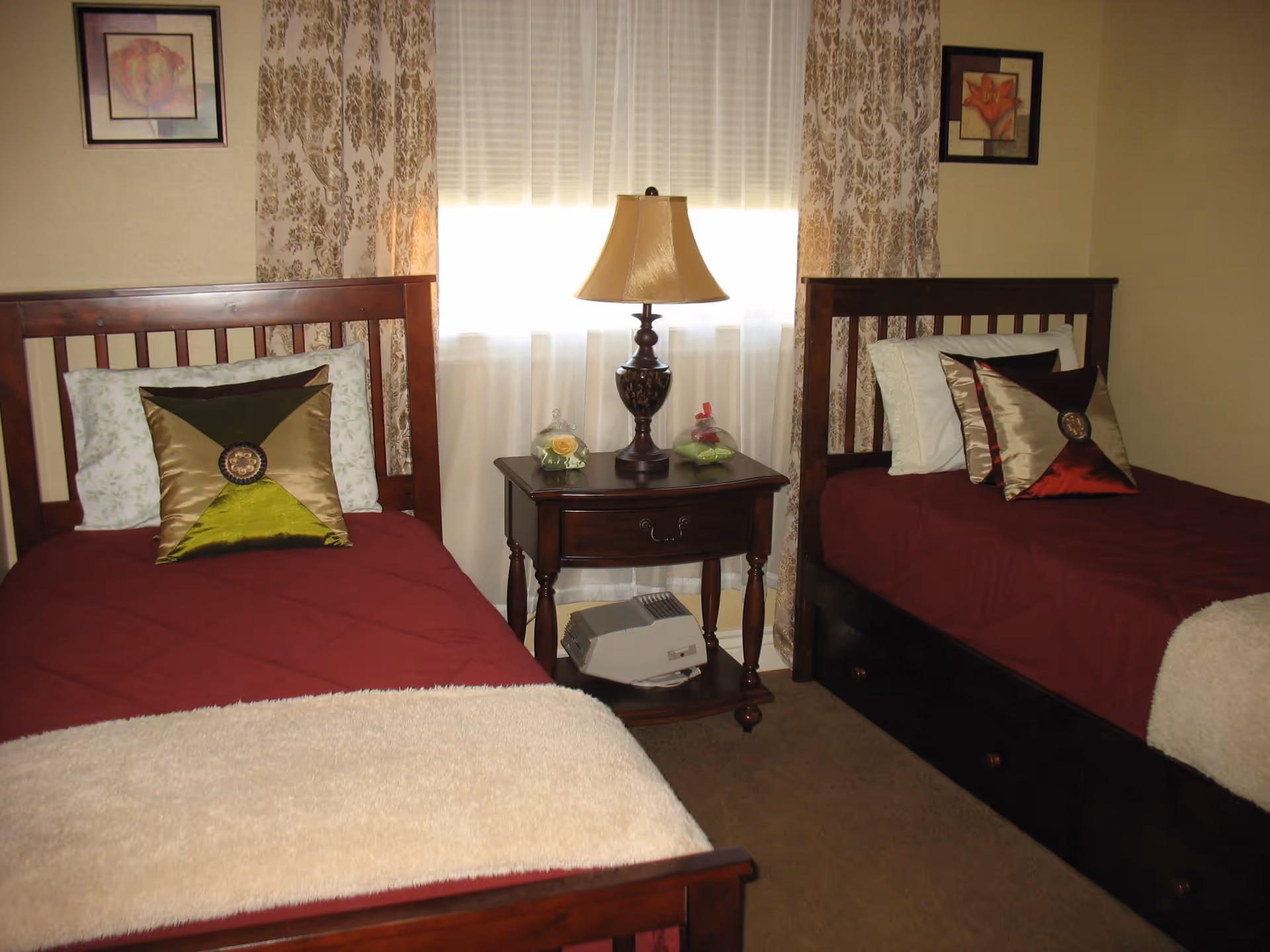 A bedroom with two single wooden beds, each with a burgundy quilt, decorative pillows, and a cream-colored throw blanket at the foot. Between the beds is a wooden nightstand with a table lamp and two small decorative items. The window behind the nightstand is covered with sheer white curtains and patterned drapes. Two framed floral pictures hang on the walls above each bed.