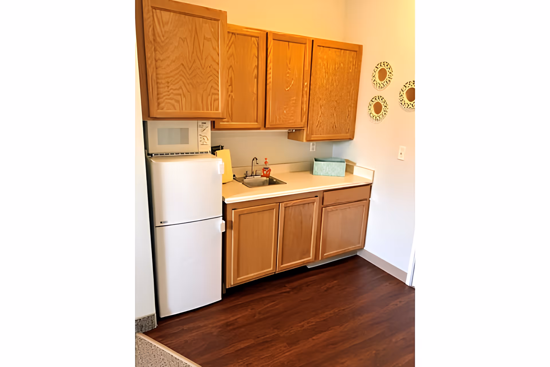 Small kitchen area with wooden cabinets, a white refrigerator, a microwave on top of the refrigerator, a countertop with a small sink, a soap dispenser, and a green tissue box. Three decorative round mirrors are mounted on the wall to the right.