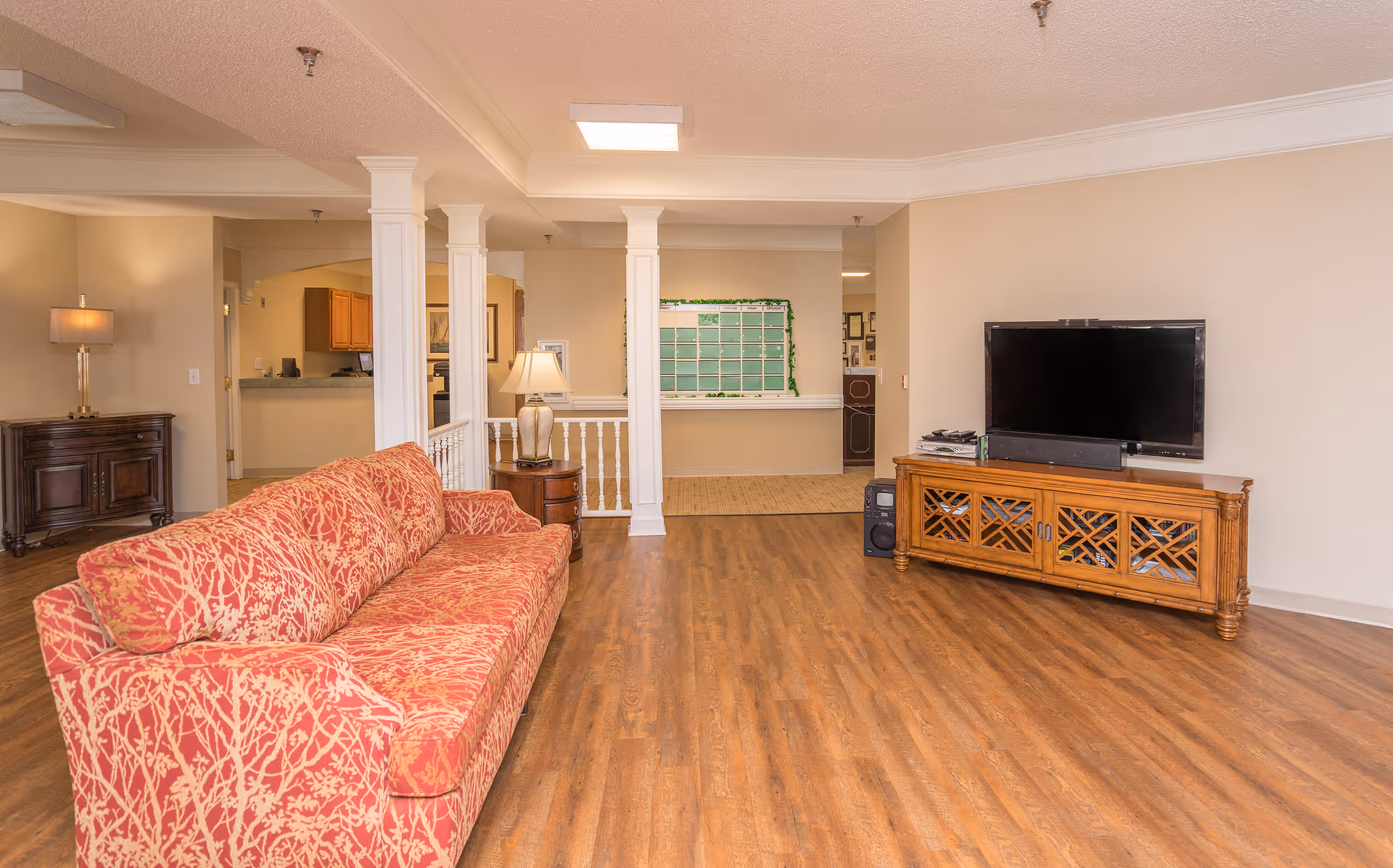 A spacious living room area with a red patterned sofa on the left, a wooden TV stand with a flat-screen television on the right, hardwood flooring, and beige walls. There are white columns and a small table with a lamp near the sofa. In the background, there is an open doorway leading to a kitchen area and a bulletin board on the far wall.