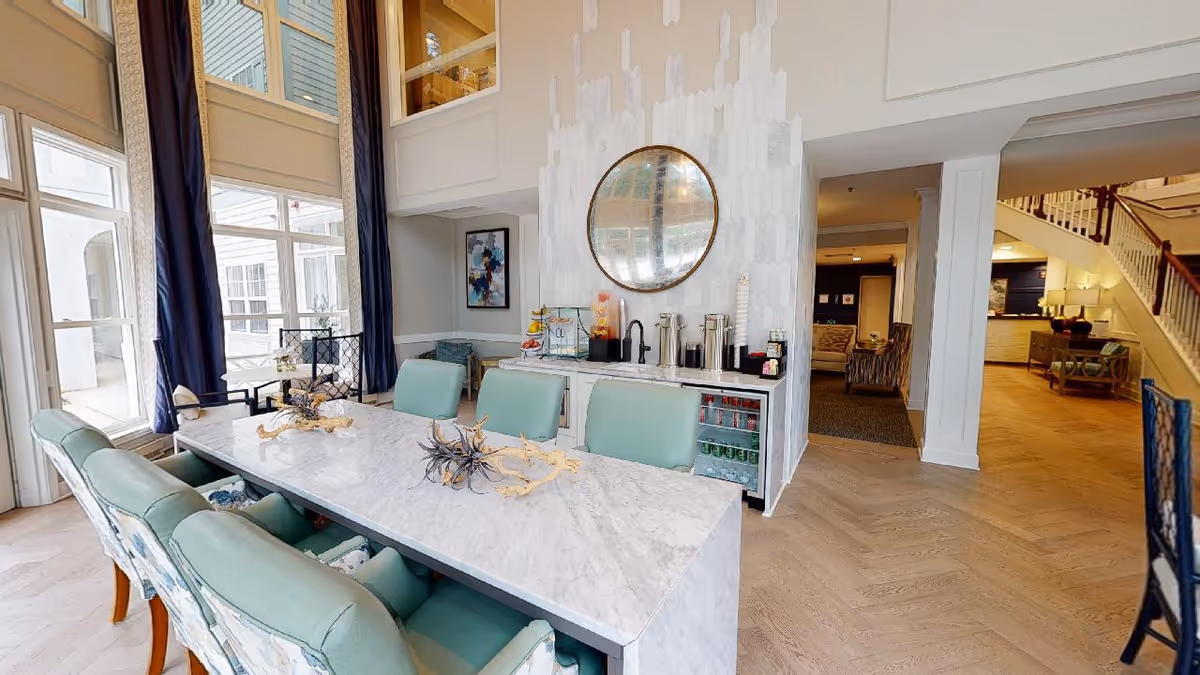 A bright and spacious interior area with a large marble table surrounded by cushioned chairs in light green and floral patterns. Behind the table is a counter with a beverage station including coffee dispensers, juice containers, and a mini fridge stocked with drinks. A large round mirror hangs on the wall above the counter. The room has tall windows with dark blue curtains, light wood flooring, and an open view into adjacent rooms and a staircase.