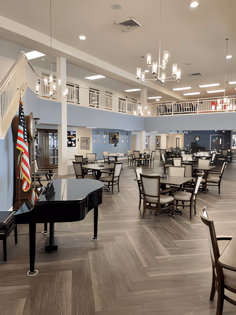 Spacious dining area with multiple tables and chairs arranged neatly. A black grand piano is positioned on the left side near a wall with an American flag. The room features high ceilings with modern chandeliers and a second-floor balcony overlooking the space.