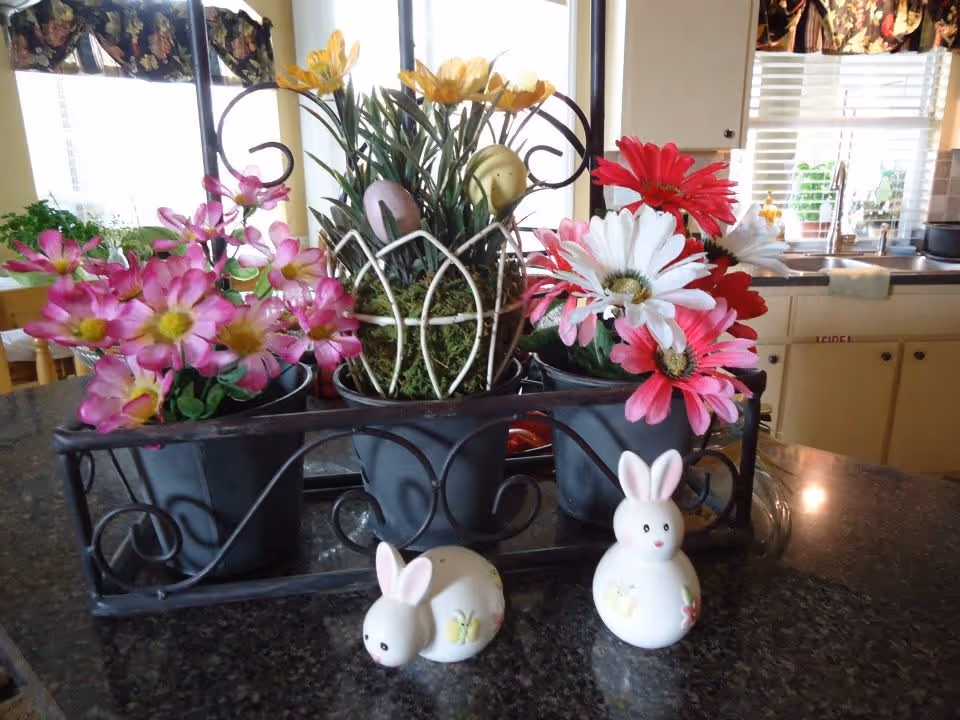 A kitchen countertop with a decorative black metal holder containing three pots of artificial flowers in pink, yellow, white, and red. In front of the pots, there are two small ceramic bunny figurines. In the background, there is a kitchen sink, cabinets, and a window with floral curtains.