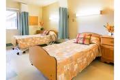 Bright shared bedroom with two hospital-style single beds, floral bedspreads, bedside table, privacy curtains and wall-mounted lights.