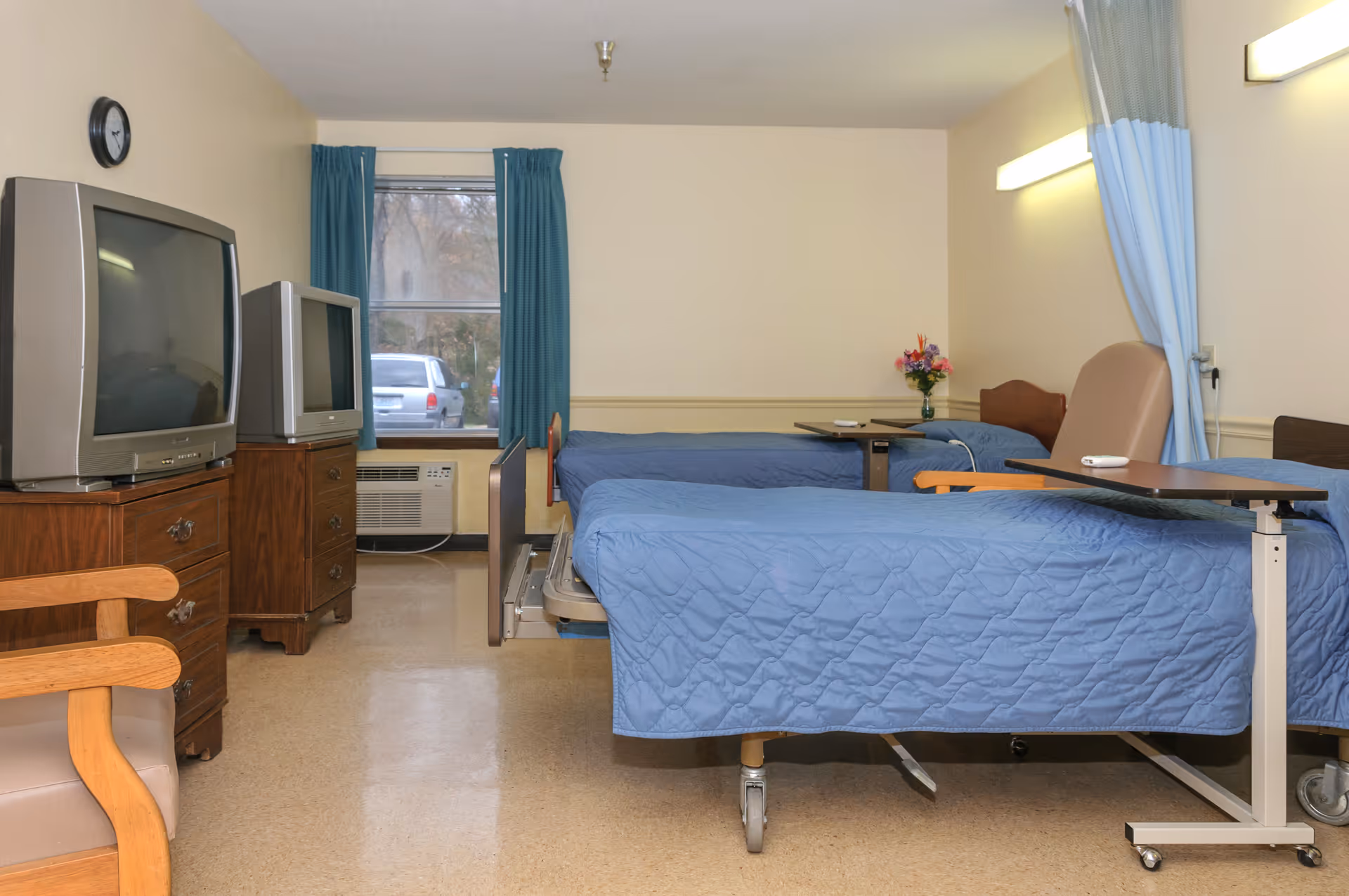 A room with two hospital-style beds covered with blue quilts, each with an overbed table. There is a wooden chair in the foreground, two older model televisions on wooden dressers, a window with blue curtains, and a small vase of flowers on a table near the back bed. The room has beige walls and flooring, with a blue privacy curtain partially visible on the right side.