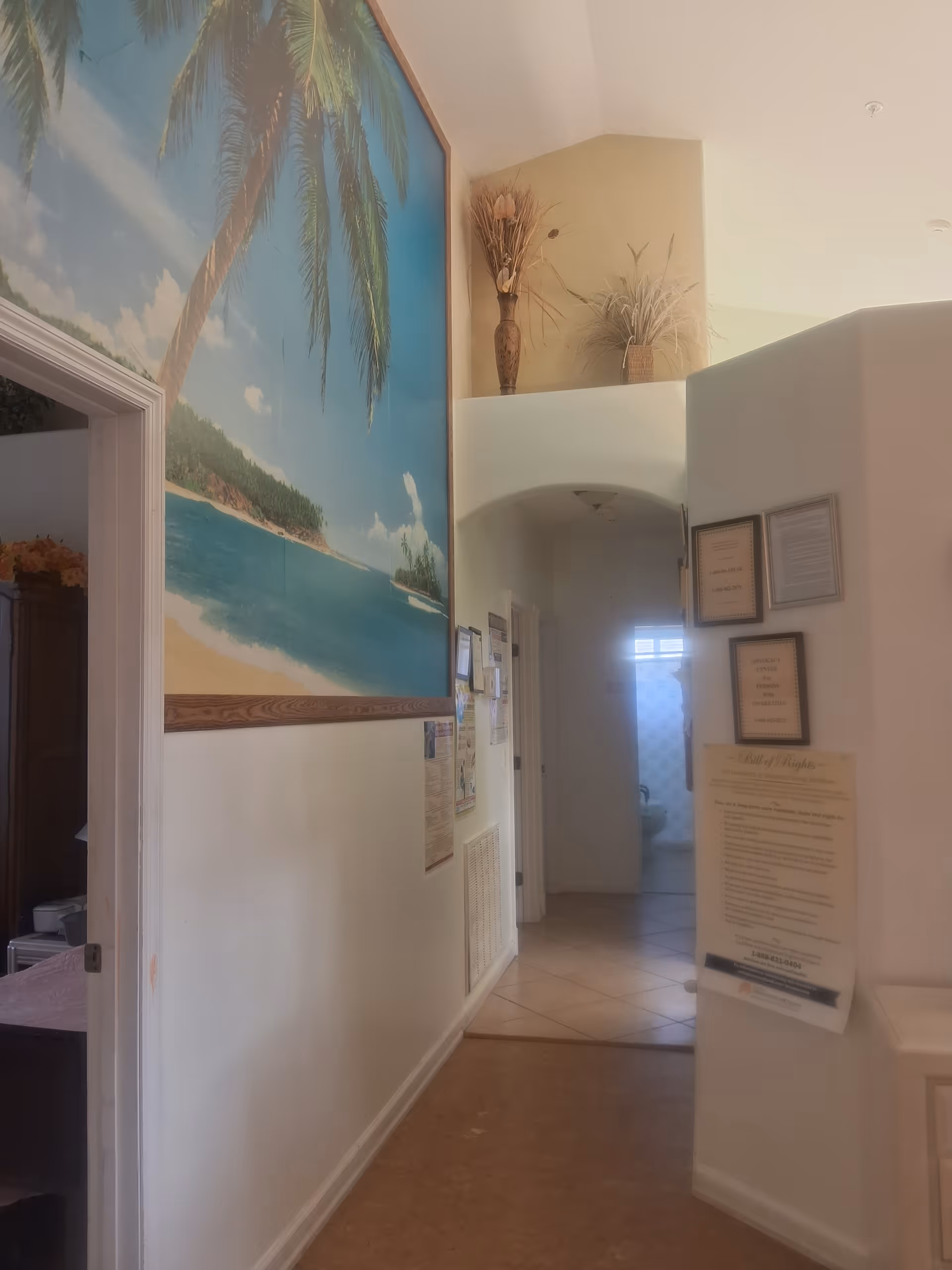 Interior hallway of Violet Gardens Assisted Living Facility with a large tropical beach mural on the left wall, decorative vases with dried plants on a ledge above an archway, and framed documents and notices on the right wall. The hallway leads to a room with a window and a partially visible toilet.