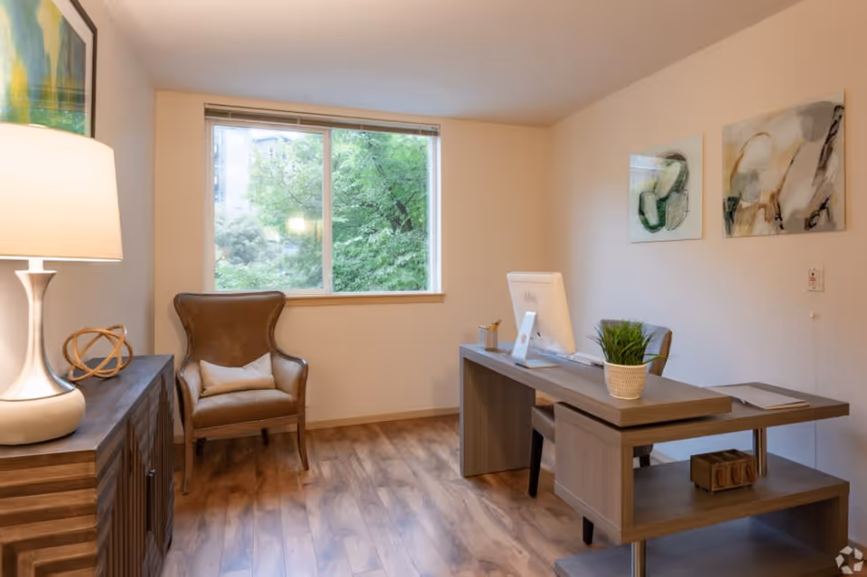 A well-lit office space with a wooden desk, a computer monitor, a potted plant, and a chair. There is a brown armchair with a pillow near a window showing green trees outside. A wooden cabinet with a lamp and decorative item is on the left side, and two abstract paintings hang on the wall.