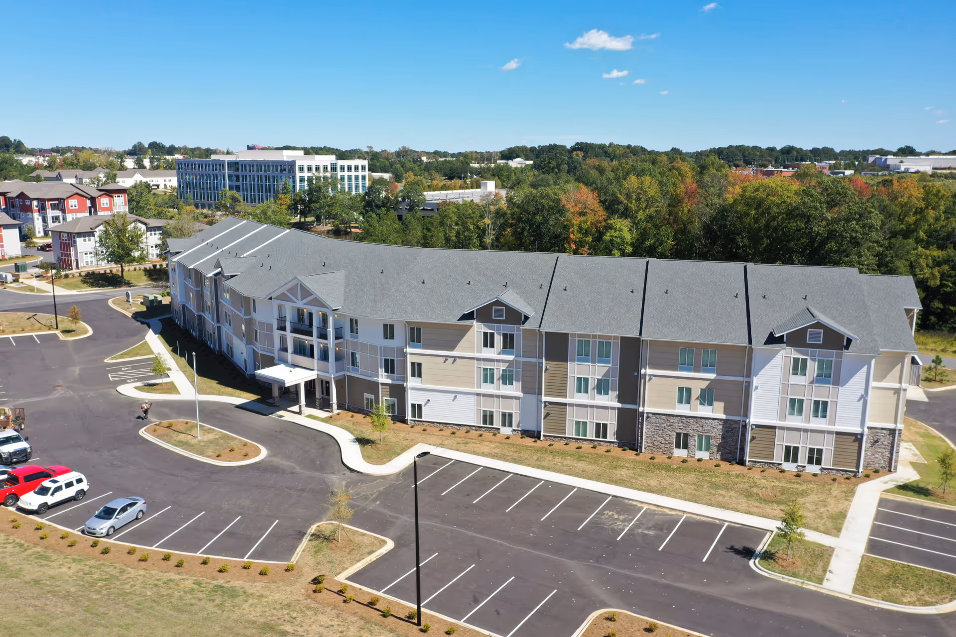 Aerial view of a large, three-story senior living facility building with a gray roof and beige and white exterior walls. The building is surrounded by parking lots with marked spaces, some cars parked, and landscaped areas with small trees and shrubs. In the background, there are other buildings and a wooded area under a clear blue sky.