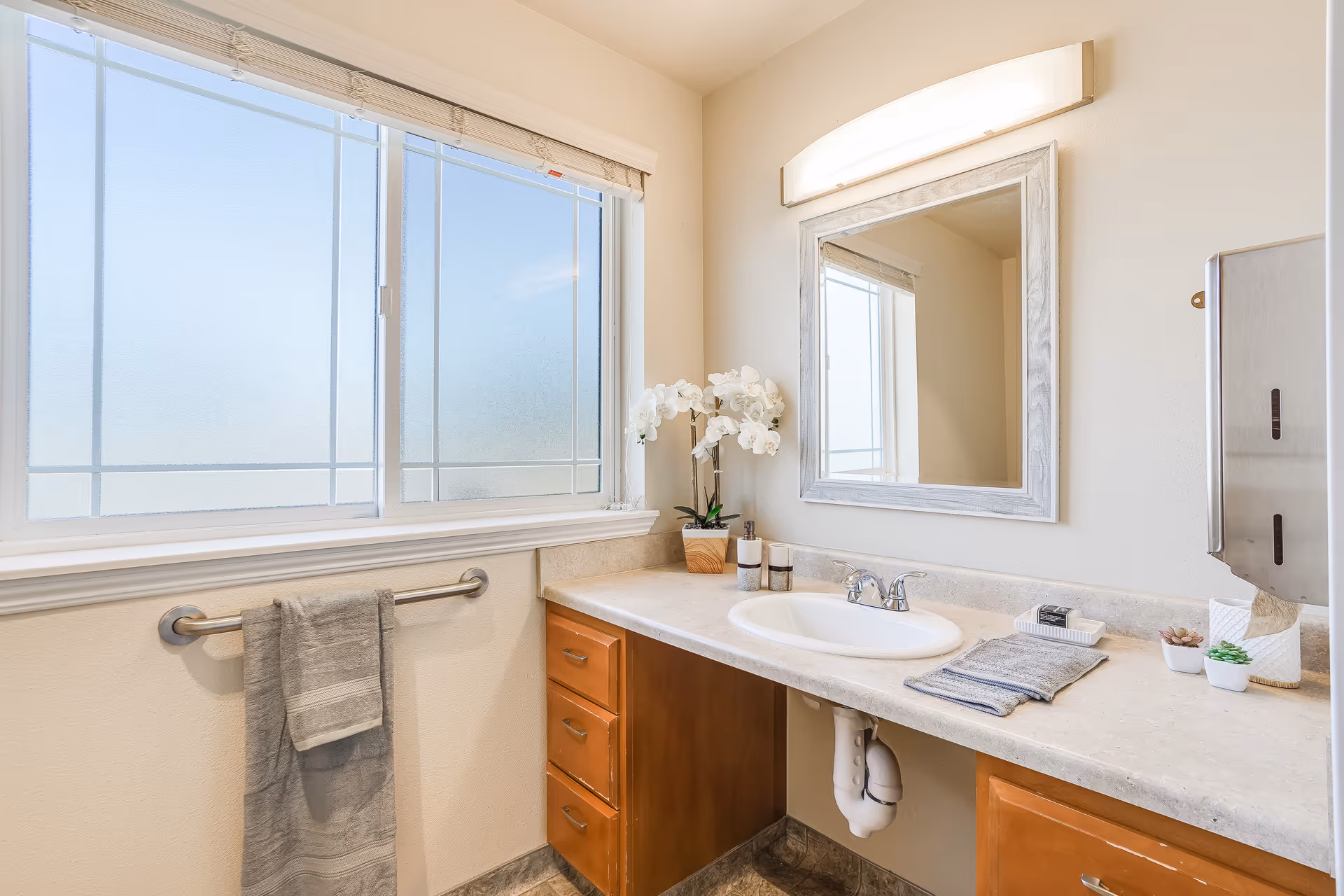 Bright bathroom with a sink and countertop beneath a mirror, a large frosted window, towel bar, and wooden cabinets.