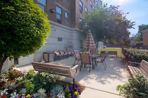 Outdoor patio area at Senior Suites Belmont Cragin with several chairs and tables, including one with a striped umbrella. There are benches, flower beds with colorful flowers, and trees providing shade. The patio is adjacent to a multi-story brick building under a clear blue sky.