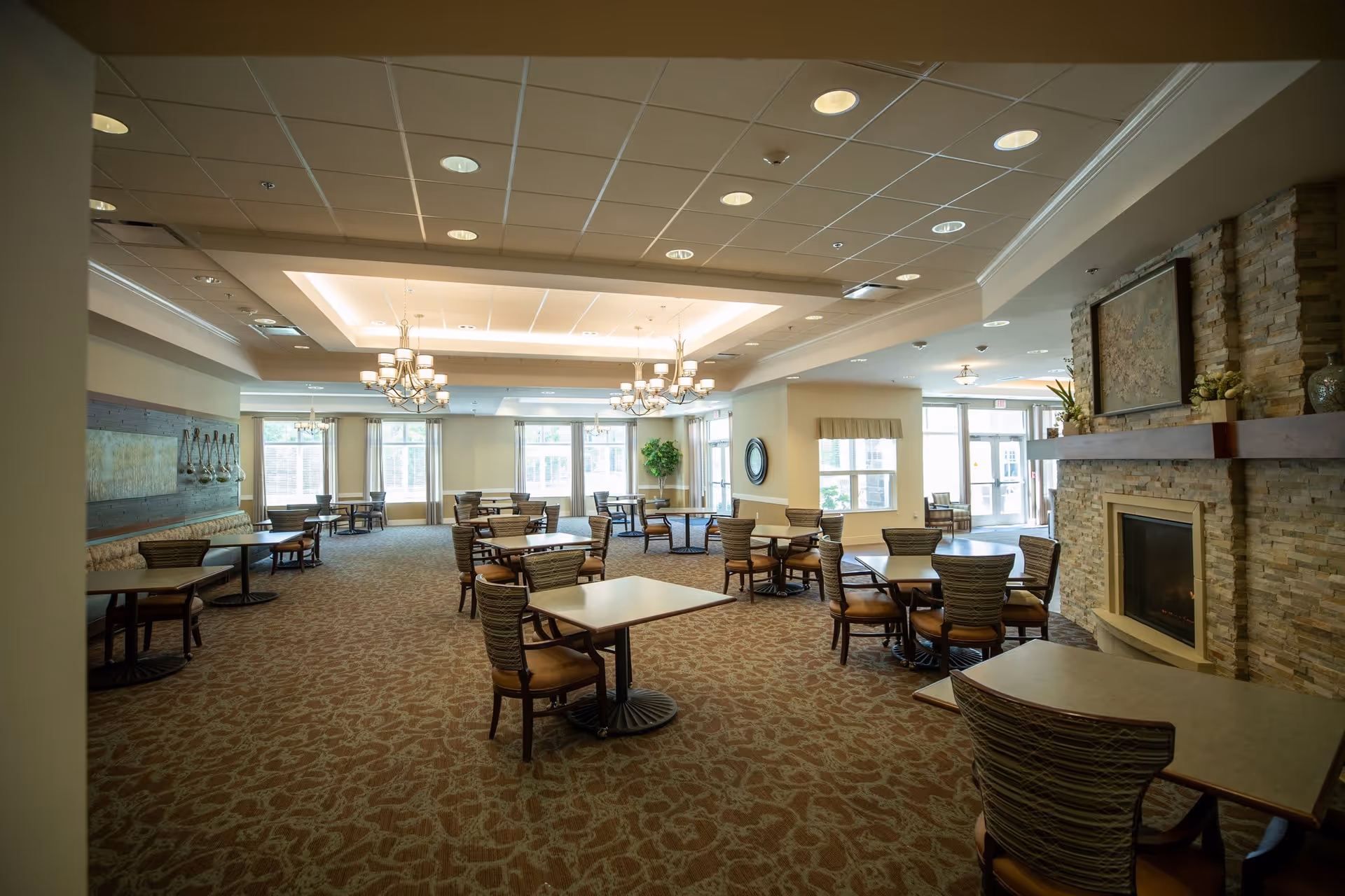 A spacious dining room with multiple square and round tables surrounded by chairs. The room features a patterned carpet, a stone fireplace with a mantel and artwork above it, large windows allowing natural light, and elegant chandeliers hanging from the ceiling.