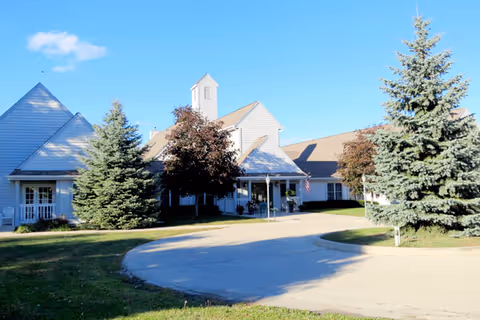 Exterior view of a single-story building with light-colored siding, a peaked roof, and a small tower-like structure. The building is surrounded by green grass, several evergreen trees, and a clear blue sky. A curved driveway leads to the entrance, which has a covered porch with an American flag.