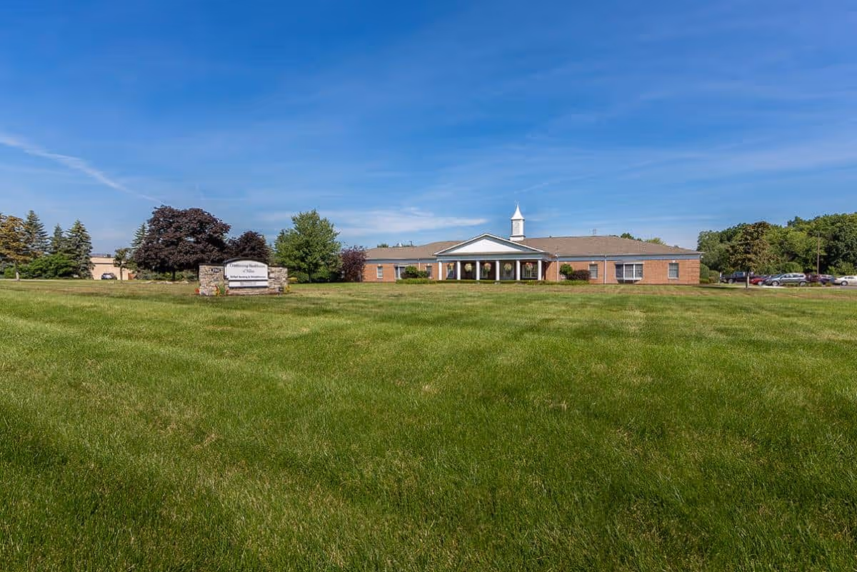 Wide view of a single-story brick building with a white cupola on the roof, surrounded by a large green lawn and trees under a clear blue sky. A stone sign in front reads 'Continuing Healthcare of Niles'.