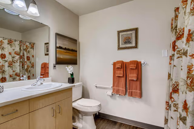A clean bathroom featuring a white toilet, a wooden vanity with a white sink, a large mirror above the sink, and a floral-patterned shower curtain. Two rust-colored towels hang on a towel rack next to the toilet, and a framed picture is mounted on the wall above the towels. The floor is a dark wood tone.