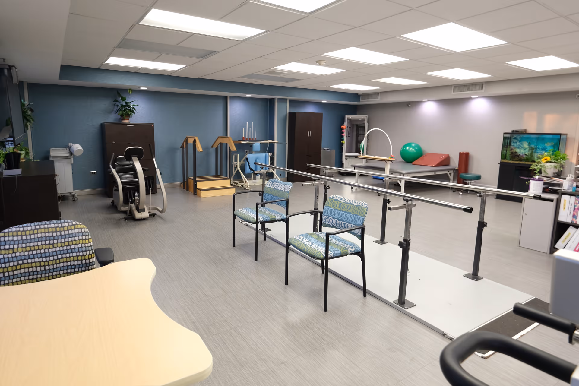 Physical therapy room with parallel bars, exercise machines, chairs, therapy tables, and an aquarium.