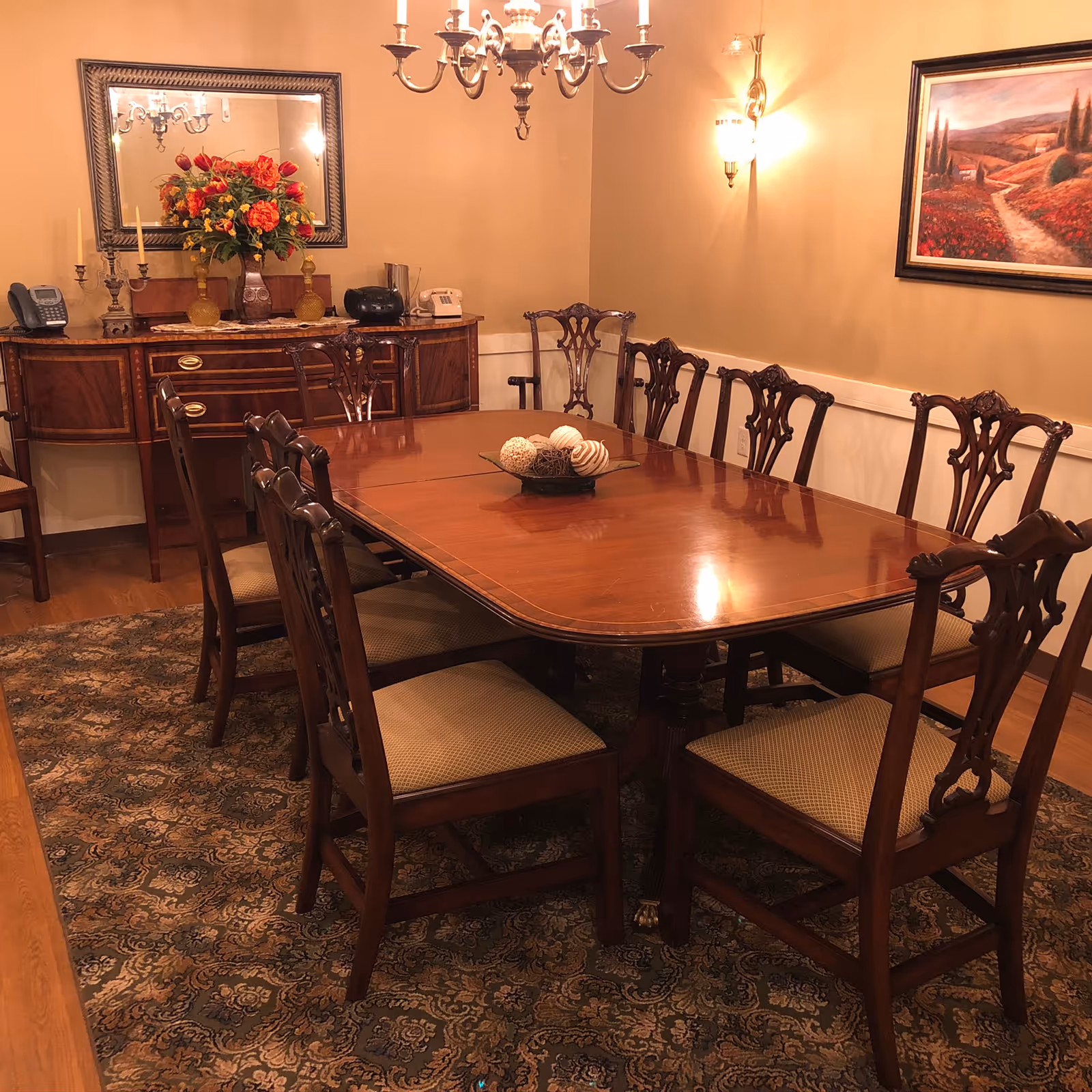 A traditional dining room with a large polished wooden dining table surrounded by eight matching wooden chairs with cushioned seats. The room features a patterned area rug, a sideboard with decorative items including a vase with flowers, candlesticks, and telephones. A chandelier hangs above the table, and a framed landscape painting is on the wall.