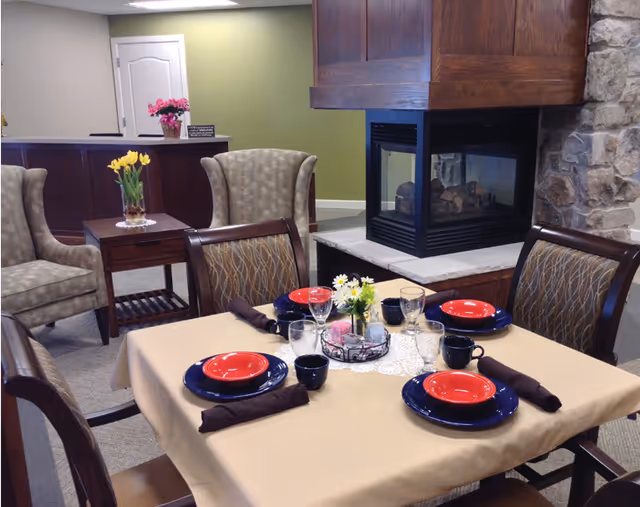 A dining area in a senior living facility with a table set for four people. The table has a beige tablecloth, blue plates with red bowls, glasses, black mugs, and brown napkins. In the center of the table is a small floral arrangement and candles. Surrounding the table are four wooden chairs with patterned upholstery. In the background, there are two upholstered armchairs, a small wooden side table with a vase of yellow flowers, and a stone fireplace with a wooden mantle. The walls are painted green and beige.