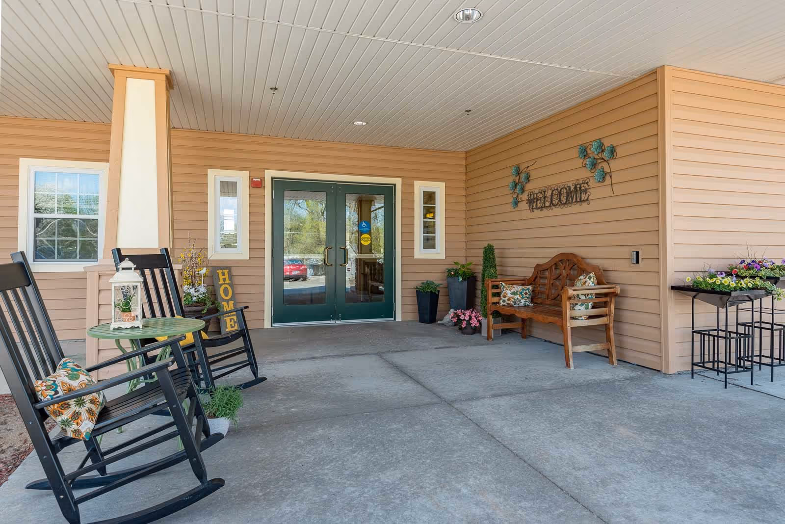 Covered entrance area of a senior living facility with double glass doors. There are two black rocking chairs with floral cushions and a small round green table with a decorative lantern on it. A wooden bench with floral pillows is placed against the wall, which has a decorative metal 'WELCOME' sign and some potted plants. The building exterior is beige with white trim around the windows and doors.