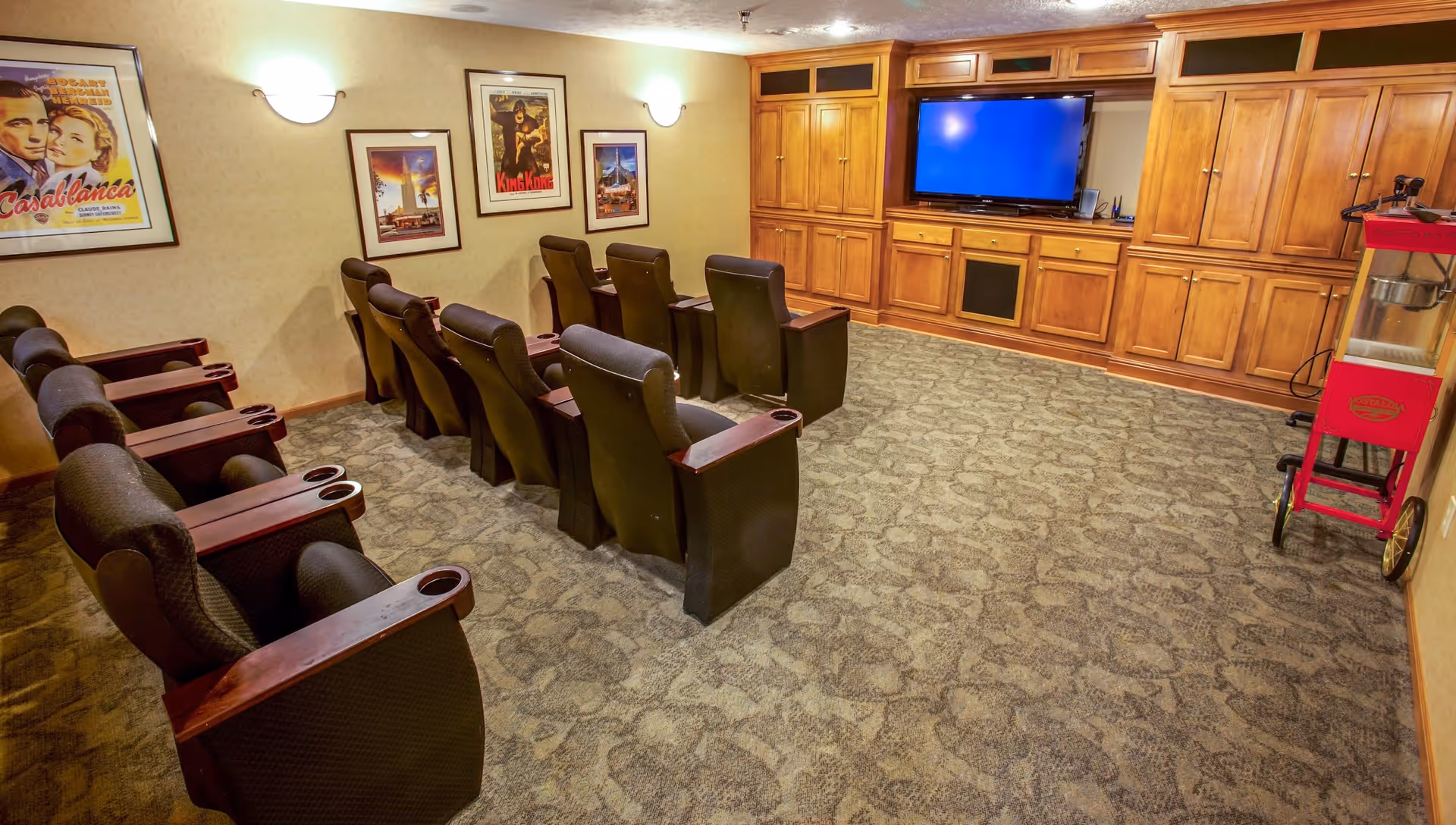 A small theater room with two rows of black cushioned chairs with wooden armrests facing a large flat-screen TV mounted on a wooden cabinet wall. The room has beige walls with three framed vintage movie posters and wall-mounted lights. A red popcorn machine is positioned on the right side of the room.