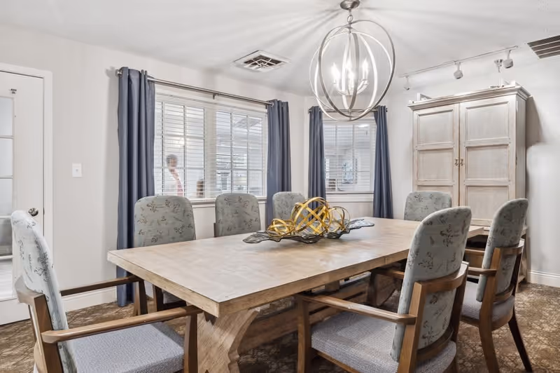 A dining room with a large wooden table surrounded by eight upholstered chairs. The table has a decorative centerpiece with golden spherical sculptures. The room features two windows with blue curtains, a white door with glass panels, a beige cabinet, and a modern spherical chandelier hanging from the ceiling.