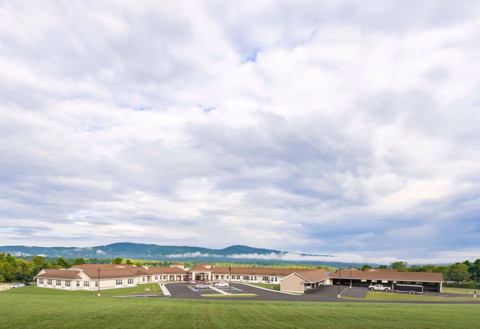 Wide exterior view of a single-story senior living facility building with a large parking area in front, surrounded by green grass and trees, with mountains and a cloudy sky in the background.