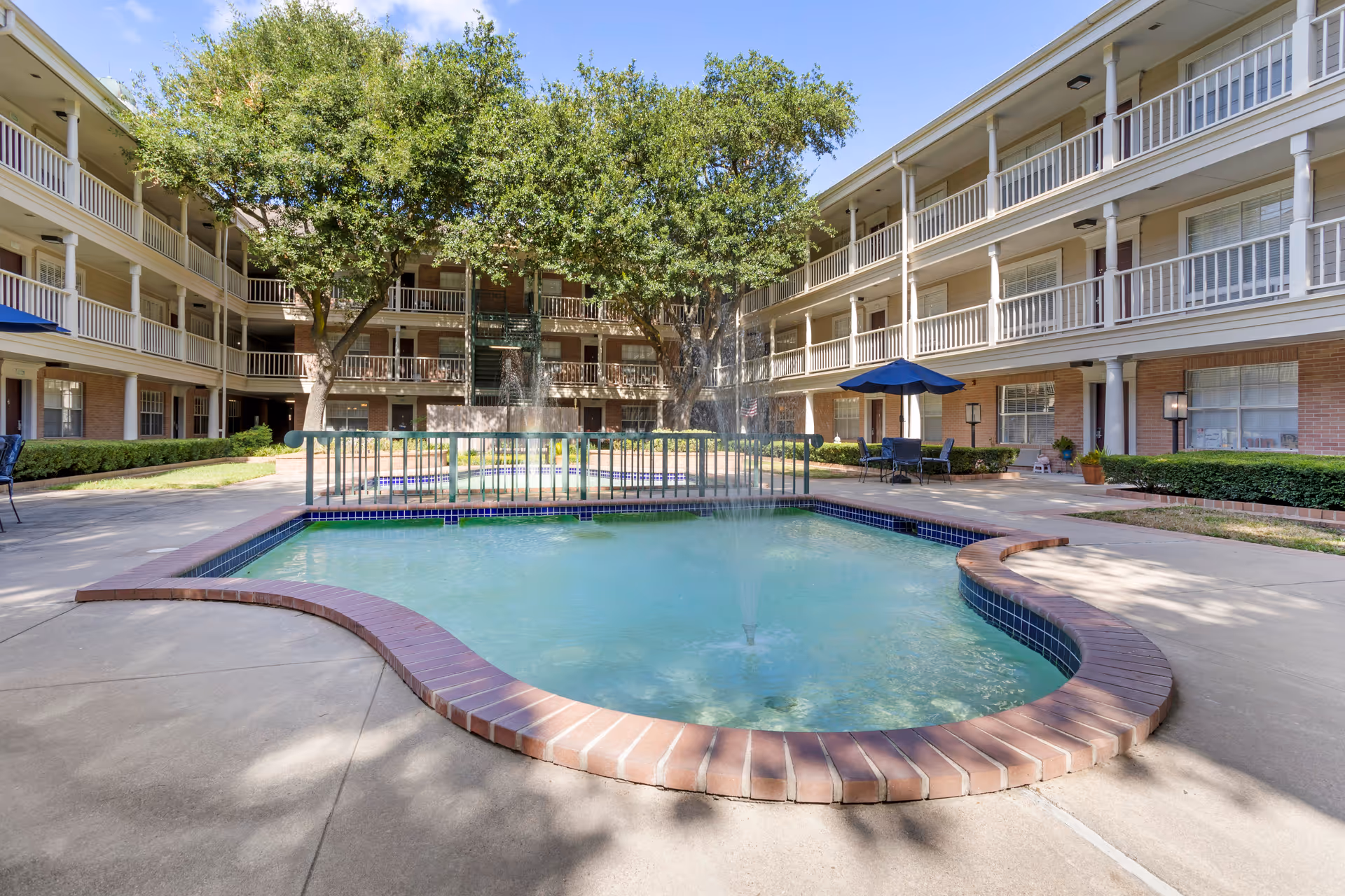 Outdoor courtyard area of a senior living facility with a small fountain pool surrounded by a brick border. The courtyard is enclosed by a three-story building with balconies and railings. There are trees providing shade and patio tables with umbrellas and chairs around the pool area.