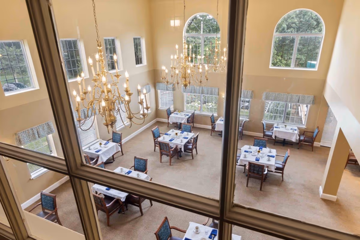 View through a window into a dining room with multiple tables set with white tablecloths, blue napkins, and dining chairs. The room features large arched windows, beige walls, and two ornate gold chandeliers hanging from a high ceiling.