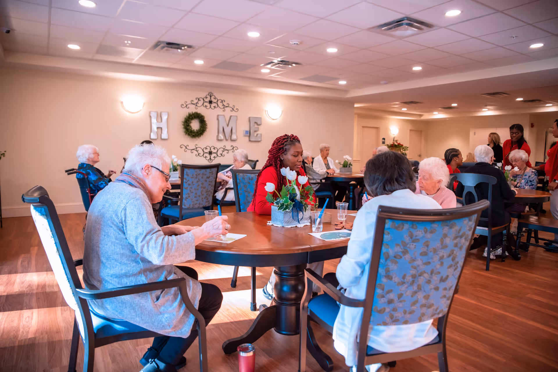 A group of elderly people and caregivers sitting around tables in a well-lit common area of a senior living facility. The room has wooden floors, decorative chairs, and a wall decoration spelling out 'HOME' with a wreath in place of the letter O. Some people are engaged in activities like writing or drawing.