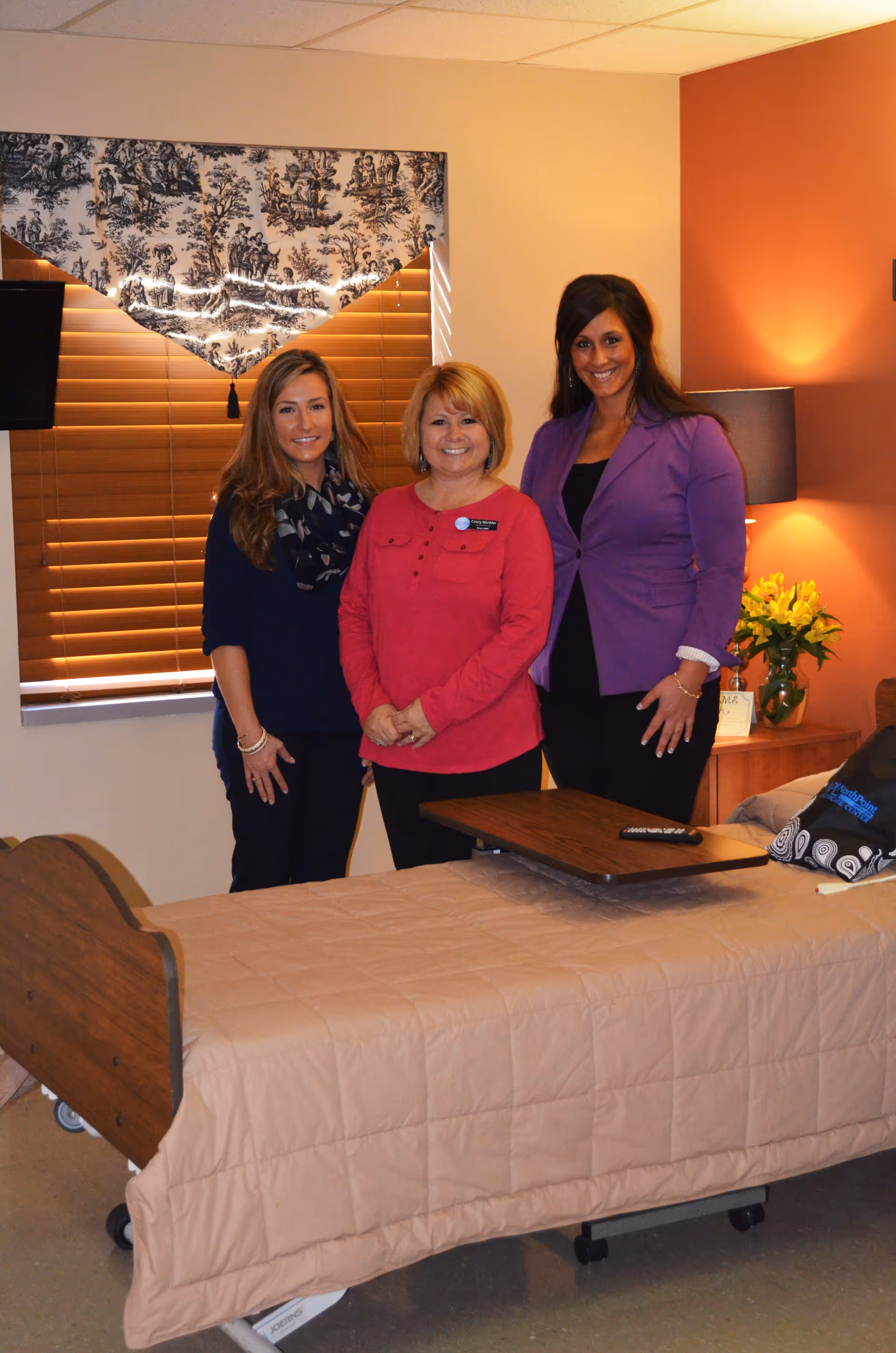 Three smiling staff members standing behind a hospital-style bed in a warmly lit resident bedroom.