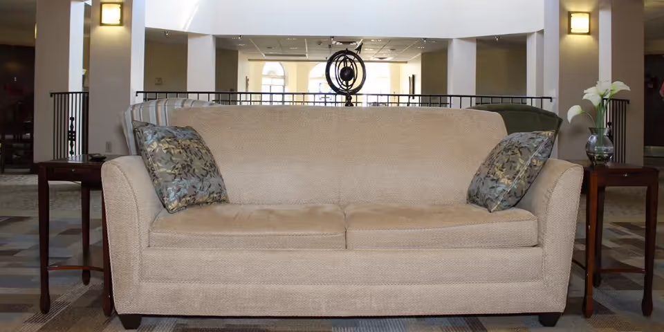 A beige upholstered sofa with two patterned throw pillows is centered in a spacious, well-lit common area. On either side of the sofa are wooden side tables, one with a glass vase holding white flowers. The background shows a railing and large windows letting in natural light.